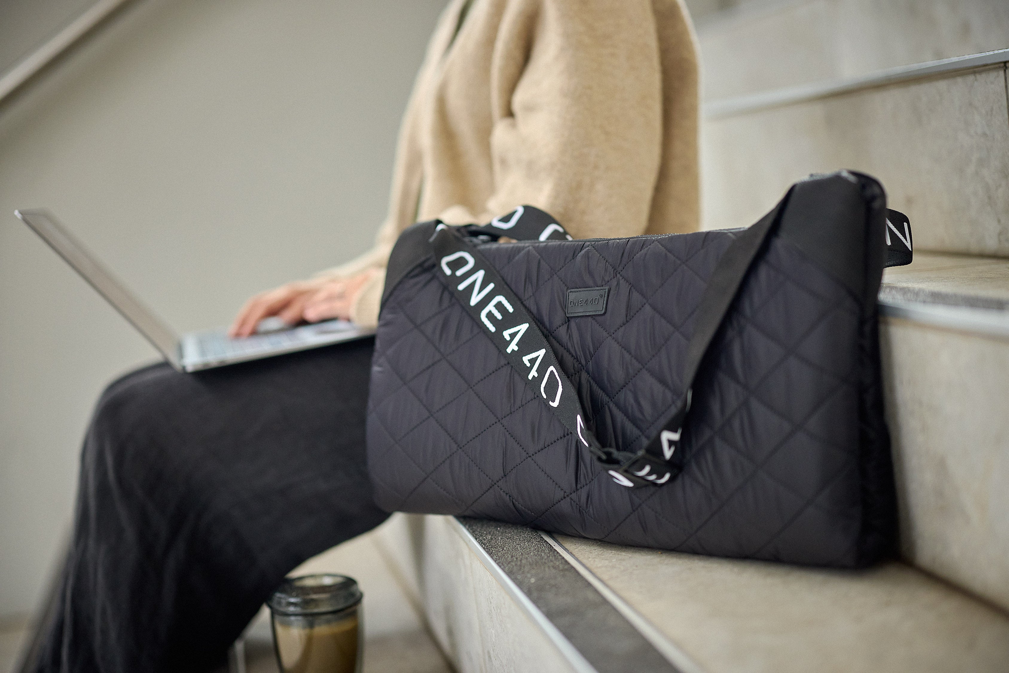 Person sitting on stairs with a black one440 quilted bag and laptop, wearing a beige coat.